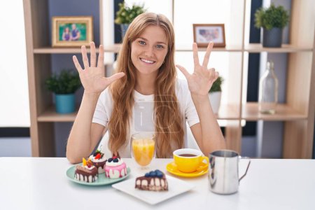 Téléchargez les photos : Jeune femme caucasienne manger pâtisseries t pour le petit déjeuner montrant et pointant vers le haut avec les doigts numéro huit tout en souriant confiant et heureux. - en image libre de droit