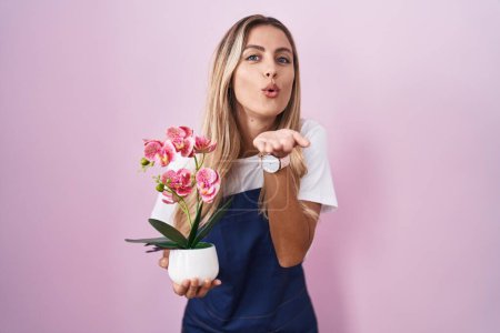 Foto de Young blonde woman wearing gardener apron holding plant looking at the camera blowing a kiss with hand on air being lovely and sexy. love expression. - Imagen libre de derechos