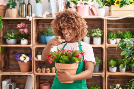 Téléchargez les photos : Jeune femme hispanique avec les cheveux bouclés travaillant à la boutique de fleuriste tenant usine souriant heureux pointant avec la main et le doigt - en image libre de droit