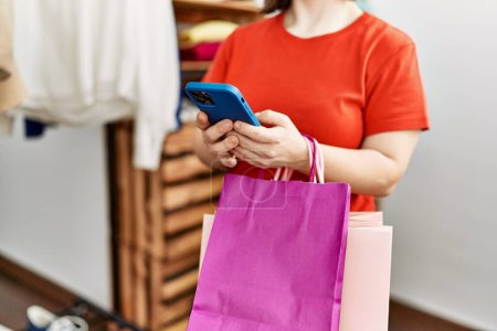 Téléchargez les photos : Femme brune avec syndrome du duvet tenant des sacs à provisions à l'aide d'un smartphone au magasin de détail - en image libre de droit