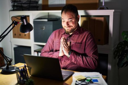 Photo for Plus size hispanic man with beard working at the office at night praying with hands together asking for forgiveness smiling confident. - Royalty Free Image