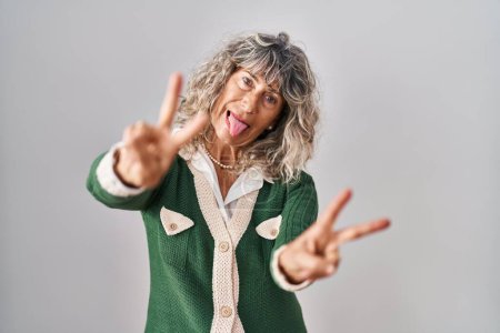 Téléchargez les photos : Middle age woman standing over white background smiling with tongue out showing fingers of both hands doing victory sign. number two. - en image libre de droit