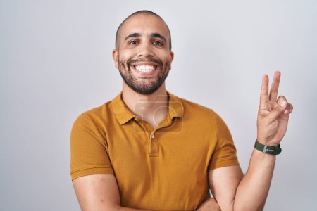 Téléchargez les photos : Hispanic man with beard standing over white background smiling with happy face winking at the camera doing victory sign with fingers. number two. - en image libre de droit