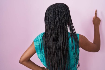 Photo for Young african american with braids standing over pink background posing backwards pointing ahead with finger hand - Royalty Free Image