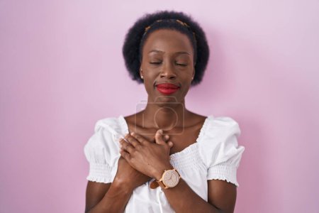 Téléchargez les photos : Femme africaine avec les cheveux bouclés debout sur fond rose souriant avec les mains sur la poitrine avec les yeux fermés et geste reconnaissant sur le visage. concept de santé. - en image libre de droit