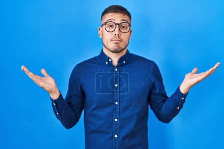 Foto de Joven hombre hispano con gafas sobre fondo azul expresión despistada y confusa con los brazos y las manos levantadas. concepto de duda. - Imagen libre de derechos