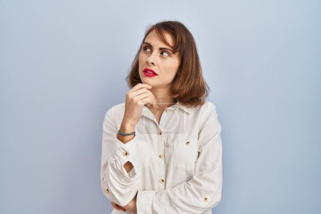 Foto de Young beautiful woman standing casual over blue background with hand on chin thinking about question, pensive expression. smiling with thoughtful face. doubt concept. - Imagen libre de derechos