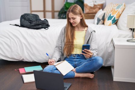 Téléchargez les photos : Young caucasian woman using smartphone and laptop writing on notebook at bedroom - en image libre de droit