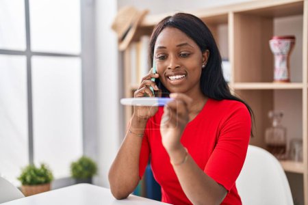 Foto de African young woman holding pregnancy test result speaking on the phone smiling with a happy and cool smile on face. showing teeth. - Imagen libre de derechos