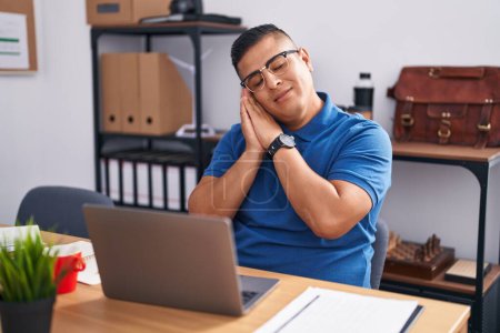 Téléchargez les photos : Jeune homme hispanique travaillant au bureau avec un ordinateur portable endormi fatigué rêvant et posant les mains ensemble tout en souriant les yeux fermés. - en image libre de droit
