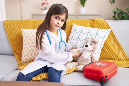 Téléchargez les photos : Adorable hispanic girl wearing doctor uniform bandaging teddy bear arm at home - en image libre de droit