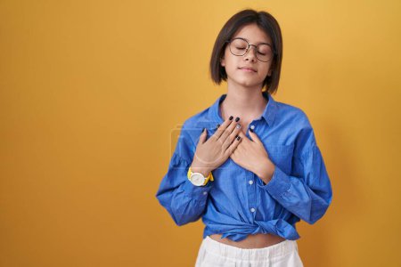 Foto de Young girl standing over yellow background smiling with hands on chest with closed eyes and grateful gesture on face. health concept. - Imagen libre de derechos