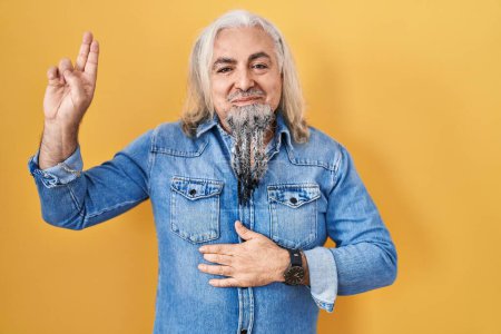 Foto de Middle age man with grey hair standing over yellow background smiling swearing with hand on chest and fingers up, making a loyalty promise oath - Imagen libre de derechos