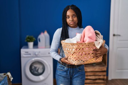 Foto de African american woman holding laundry basket thinking attitude and sober expression looking self confident - Imagen libre de derechos