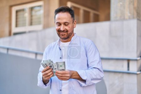Foto de Joven caucásico sonriendo confiado contando dólares en la calle - Imagen libre de derechos