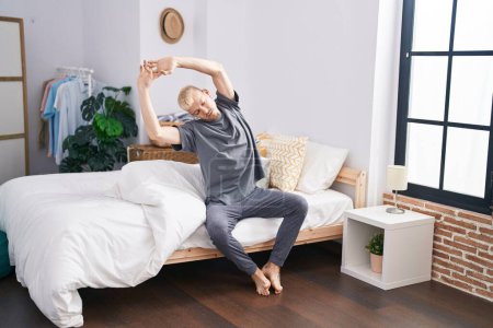 Photo for Young caucasian man stretching arms sitting on bed at bedroom - Royalty Free Image