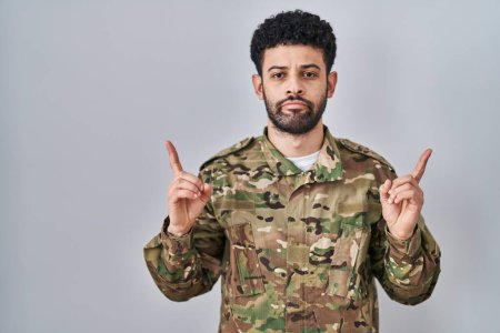 Foto de Arab man wearing camouflage army uniform pointing up looking sad and upset, indicating direction with fingers, unhappy and depressed. - Imagen libre de derechos