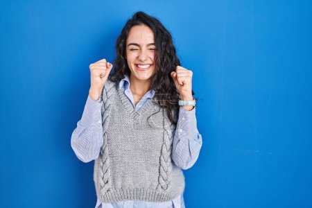 Foto de Young brunette woman standing over blue background excited for success with arms raised and eyes closed celebrating victory smiling. winner concept. - Imagen libre de derechos