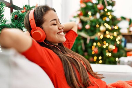 Foto de Mujer latina joven escuchando música sentada junto al árbol de Navidad en casa - Imagen libre de derechos