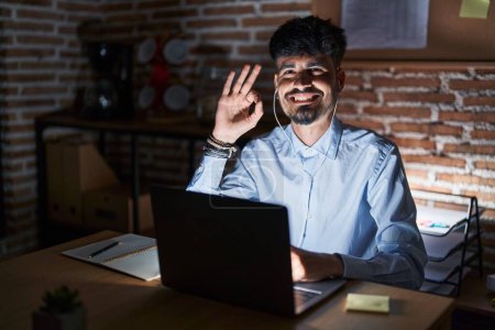 Téléchargez les photos : Young hispanic man with beard working at the office at night smiling positive doing ok sign with hand and fingers. successful expression. - en image libre de droit