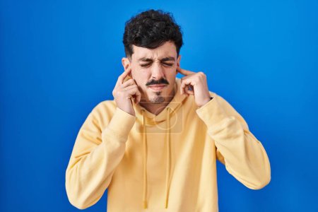 Téléchargez les photos : Homme hispanique debout sur fond bleu couvrant les oreilles avec des doigts avec une expression agacée pour le bruit de la musique forte. concept sourd. - en image libre de droit