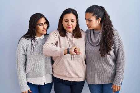 Photo for Mother and two daughters standing over blue background checking the time on wrist watch, relaxed and confident - Royalty Free Image