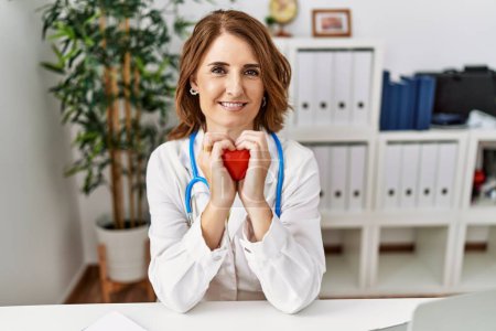 Photo for Middle age hispanic woman wearing doctor uniform holding heart at clinic - Royalty Free Image
