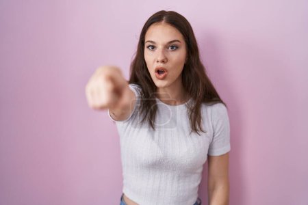 Téléchargez les photos : Young hispanic girl standing over pink background pointing with finger surprised ahead, open mouth amazed expression, something on the front - en image libre de droit