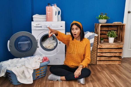 Photo for Young hispanic woman doing laundry pointing with finger surprised ahead, open mouth amazed expression, something on the front - Royalty Free Image