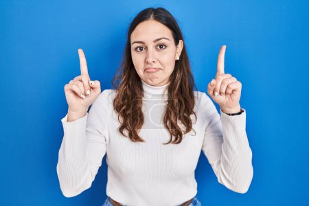 Photo for Young hispanic woman standing over blue background pointing up looking sad and upset, indicating direction with fingers, unhappy and depressed. - Royalty Free Image