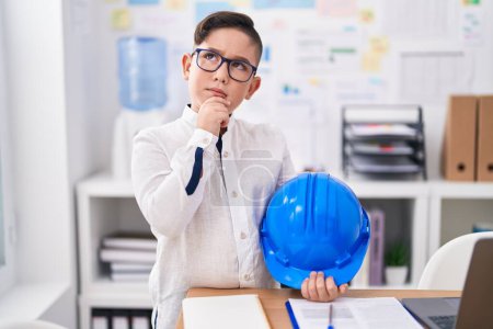 Photo for Young hispanic kid holding architect hardhat at the office serious face thinking about question with hand on chin, thoughtful about confusing idea - Royalty Free Image