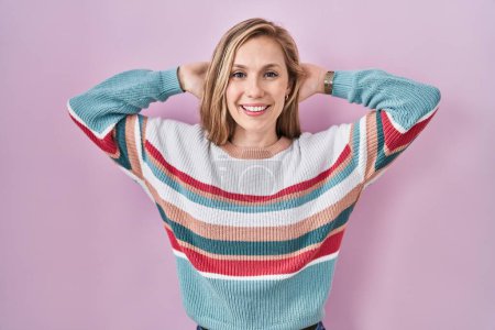 Foto de Young blonde woman standing over pink background relaxing and stretching, arms and hands behind head and neck smiling happy - Imagen libre de derechos