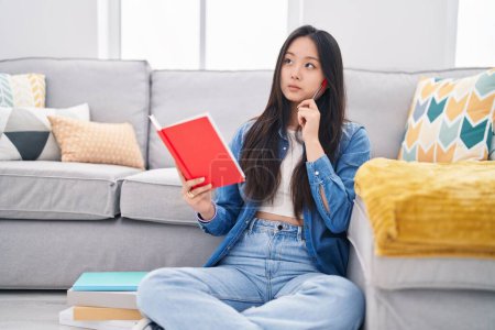 Téléchargez les photos : Jeune femme chinoise lecture livre avec l'expression du doute à la maison - en image libre de droit