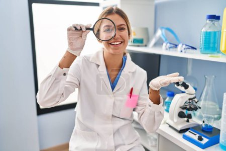 Foto de Mujer rubia joven que trabaja en el laboratorio científico usando lupa celebrando el logro con sonrisa feliz y expresión ganadora con la mano levantada - Imagen libre de derechos