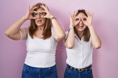 Photo for Hispanic mother and daughter wearing casual white t shirt over pink background doing ok gesture like binoculars sticking tongue out, eyes looking through fingers. crazy expression. - Royalty Free Image