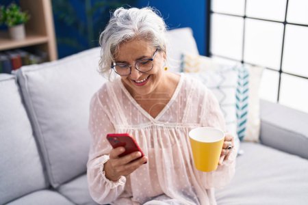 Foto de Mujer de pelo gris de mediana edad usando teléfono inteligente bebiendo café en casa - Imagen libre de derechos
