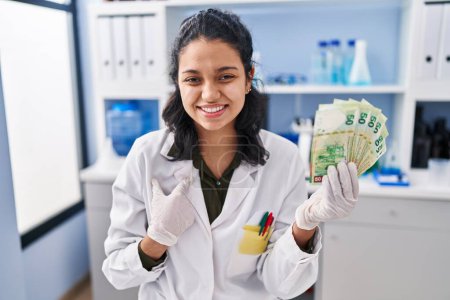 Photo for Hispanic woman with dark hair working at scientist laboratory holding money pointing finger to one self smiling happy and proud - Royalty Free Image