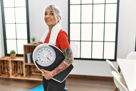 Téléchargez les photos : Moyen âge femme aux cheveux gris souriant confiant tenant machine de pesage à la maison - en image libre de droit