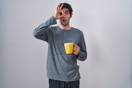 Téléchargez les photos : Jeune homme hispanique en pyjama buvant une tasse de café faisant un geste correct choqué par le visage surpris, les yeux regardant à travers les doigts. expression incrédule. - en image libre de droit