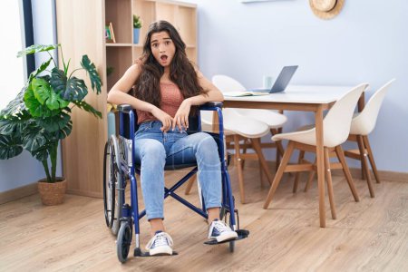 Téléchargez les photos : Young teenager girl sitting on wheelchair at the living room scared and amazed with open mouth for surprise, disbelief face - en image libre de droit