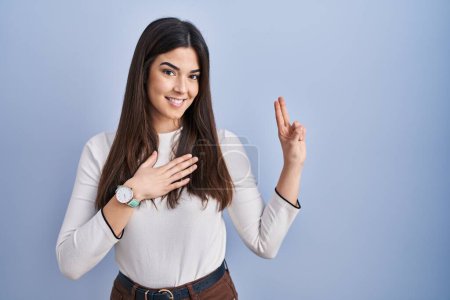 Foto de Young brunette woman standing over blue background smiling swearing with hand on chest and fingers up, making a loyalty promise oath - Imagen libre de derechos