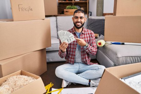 Téléchargez les photos : Middle east man with beard sitting on the floor at new home holding money smiling happy pointing with hand and finger - en image libre de droit