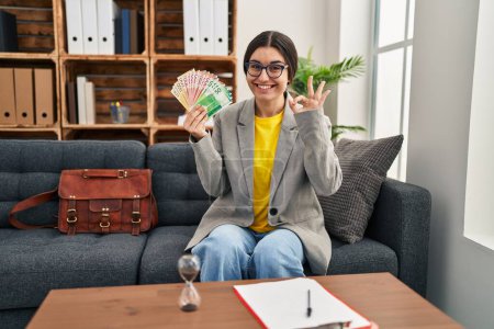 Photo for Young hispanic woman working at consultation office holding money doing ok sign with fingers, smiling friendly gesturing excellent symbol - Royalty Free Image
