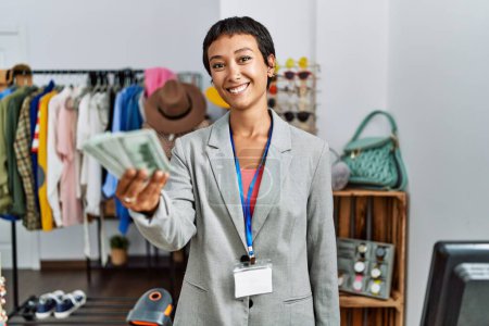 Téléchargez les photos : Jeune commerçante hispanique souriante confiante tenant des billets en dollars au magasin de vêtements - en image libre de droit