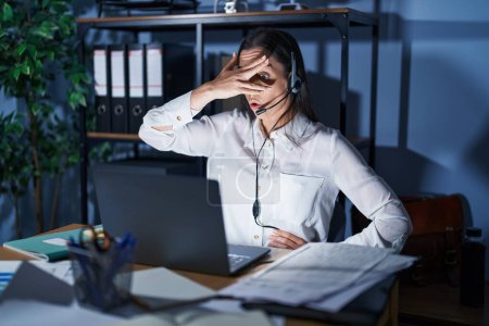 Photo for Young brunette woman wearing call center agent headset working late at night peeking in shock covering face and eyes with hand, looking through fingers with embarrassed expression. - Royalty Free Image