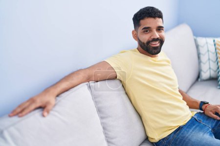 Téléchargez les photos : Jeune homme arabe souriant confiant assis sur le canapé à la maison - en image libre de droit