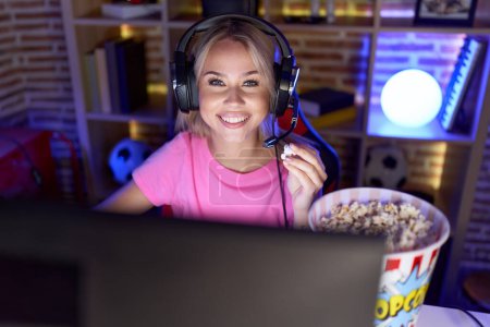 Téléchargez les photos : Jeune femme blonde streamer jouer jeu vidéo manger pop-corn à la salle de jeux - en image libre de droit
