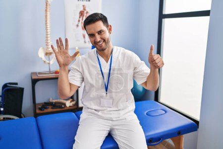Téléchargez les photos : Young hispanic man with beard working at pain recovery clinic showing and pointing up with fingers number six while smiling confident and happy. - en image libre de droit