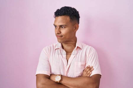 Téléchargez les photos : Jeune homme hispanique debout sur fond rose regardant du côté avec les bras croisés convaincu et confiant - en image libre de droit
