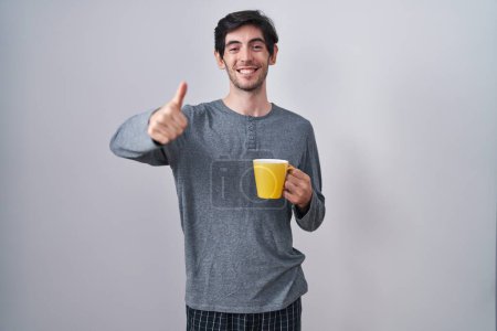 Foto de Young hispanic man wearing pajama drinking a cup of coffee approving doing positive gesture with hand, thumbs up smiling and happy for success. winner gesture. - Imagen libre de derechos